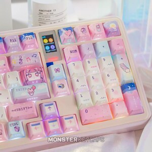 May include: A mechanical keyboard featuring a pastel color palette of pink, blue, and white. The keys display various icons including hearts, musical notes, a rabbit, and a small screen. The text "MONSTERKEYCAPS" is printed at the bottom edge.