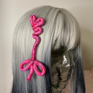 May include: A bright pink hair accessory featuring a heart-shaped top connected to a long, winding stem that ends in a bow. The surface has a glossy, textured finish with subtle purple and blue highlights, attached to a silver-gray wig.