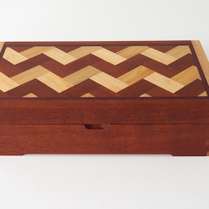 May include: A rectangular wooden box with a chevron pattern on the lid. The box is made of reddish-brown wood with lighter wood accents. The lid features a geometric design in shades of brown and tan. The box has a small recessed handle on the front.