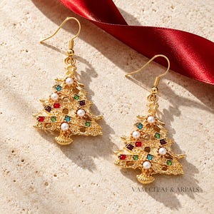 Puede incluir: Un par de pendientes colgantes dorados con forma de árbol de Navidad, decorados con pequeñas perlas blancas de imitación y cristales incrustados en rojo, verde y morado. Cuelgan de ganchos dorados e incluyen el texto VAM CLEAF & ARPALS.