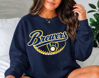 May include: A navy blue crewneck sweatshirt featuring the word Brewers in a stylized blue and yellow font, accompanied by a graphic of a baseball glove holding a baseball with yellow stitching details across the chest area.