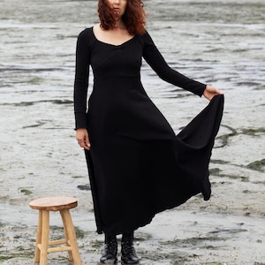 Pu&ograve; includere: Una donna con un vestito nero si trova su una spiaggia con uno sgabello di legno di fronte a lei. La spiaggia &egrave; ricoperta di alghe e il cielo &egrave; nuvoloso.