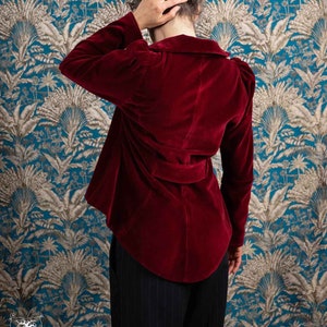 Peut inclure: Une femme portant un blazer en velours rouge avec une ceinture. Le blazer a un col et des manches longues. La femme est debout devant un papier peint à motifs.