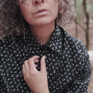 Può includere: Una donna con i capelli ricci grigi indossa una camicia a pois neri e bianchi. È in piedi davanti a uno sfondo sfocato di alberi.