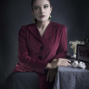 May include: A woman wearing a burgundy velvet jacket with a black skirt and green earrings. She is sitting at a table with a white tablecloth and a small framed picture with a butterfly design.