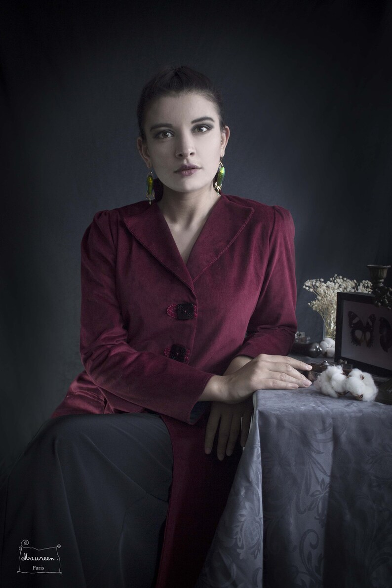 May include: A woman wearing a burgundy velvet jacket with a black skirt and green earrings. She is sitting at a table with a white tablecloth and a small framed picture with a butterfly design.