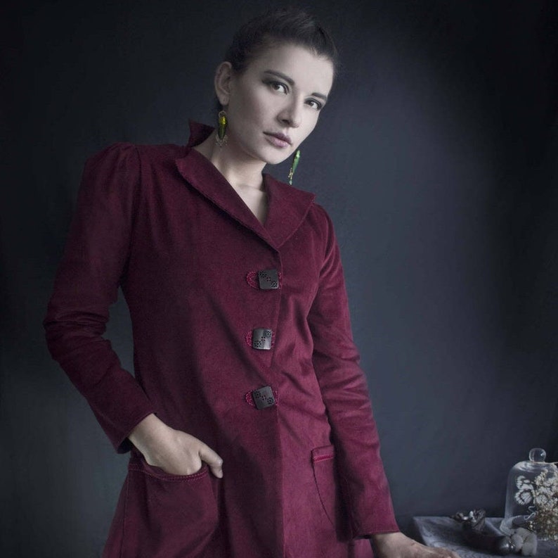 May include: A woman wearing a maroon corduroy jacket with decorative buttons. The jacket has a collar and long sleeves. She is wearing green earrings.