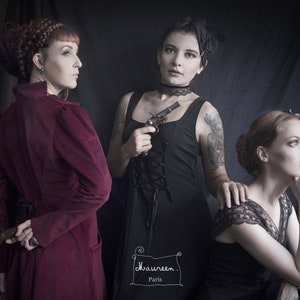 May include: Three women in a dimly lit room, wearing vintage clothing. The woman in the center is holding a small black pistol. The woman on the left is wearing a burgundy velvet coat and holding a pistol behind her back. The woman on the right is wearing a black lace top and has her hand on her chin. The text "Maureen. Paris" is written in the lower center of the image.