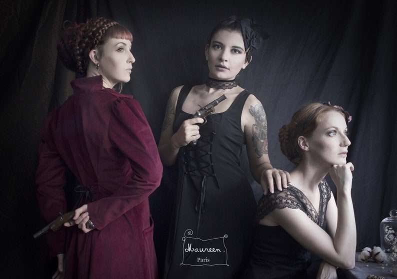 May include: Three women in a dimly lit room, wearing vintage clothing. The woman in the center is holding a small black pistol. The woman on the left is wearing a burgundy velvet coat and holding a pistol behind her back. The woman on the right is wearing a black lace top and has her hand on her chin. The text "Maureen. Paris" is written in the lower center of the image.