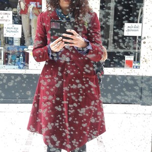 May include: A woman wearing a red velvet coat with a black phone in her hand. The coat has a pattern of small white dots. The woman is standing in front of a window with a blurry background.