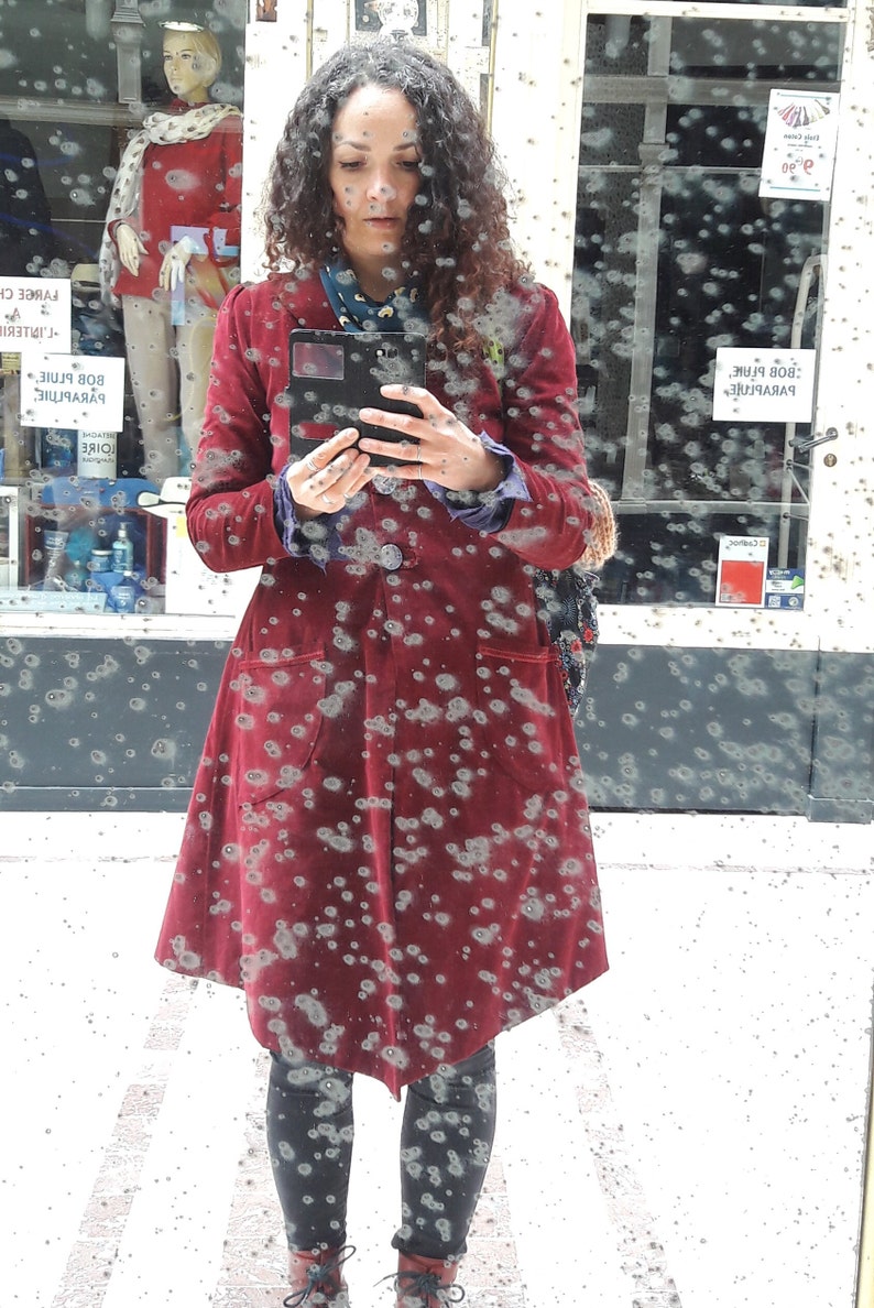 May include: A woman wearing a red velvet coat with a black phone in her hand. The coat has a pattern of small white dots. The woman is standing in front of a window with a blurry background.