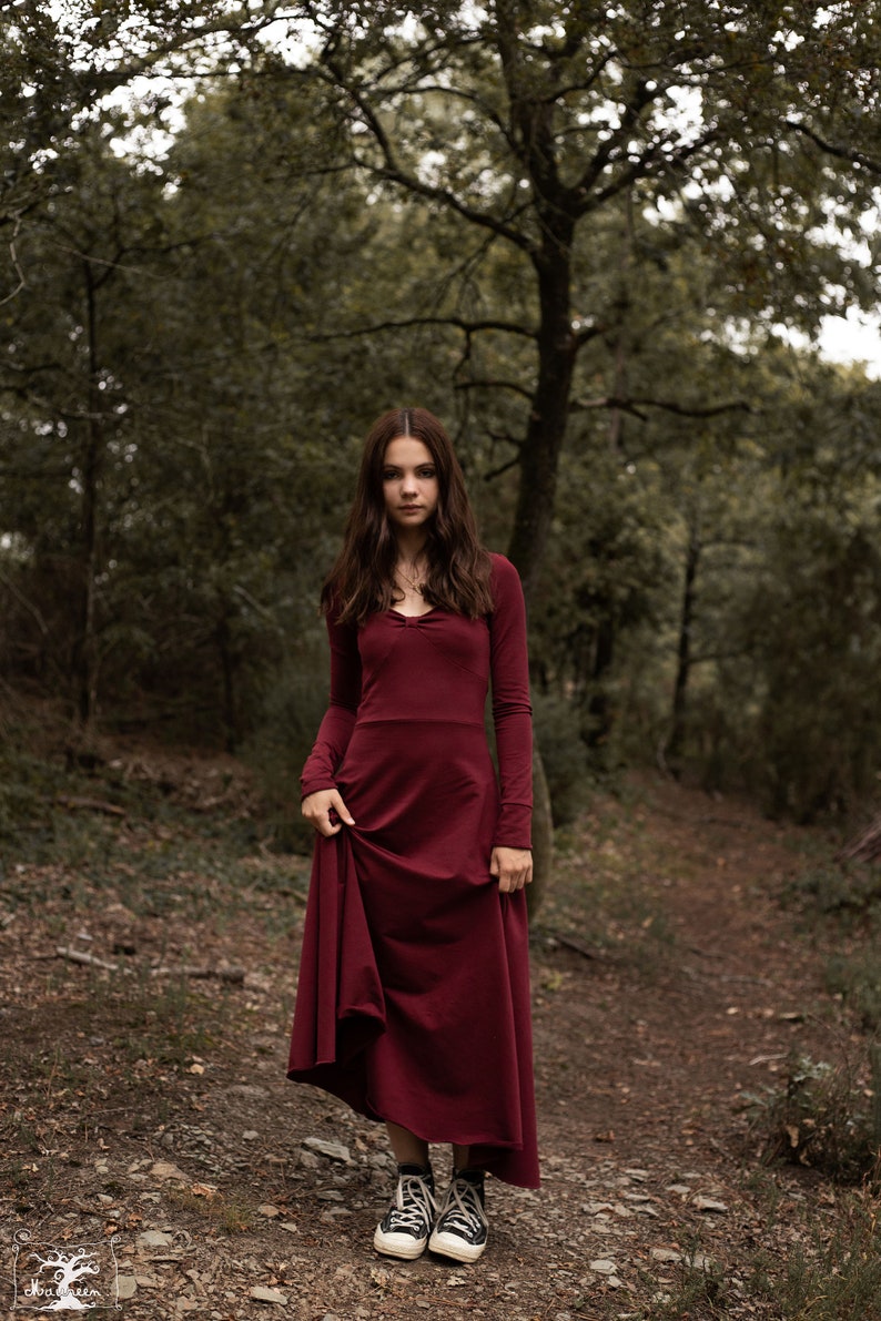 May include: A young person wearing a long, burgundy dress with long sleeves and a gathered neckline. They are standing in a forest setting on a dirt path, wearing black and white sneakers.