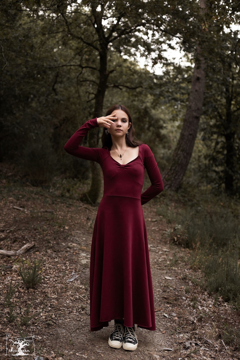 May include: A woman wearing a long, burgundy dress with a sweetheart neckline stands in a forest setting. She is wearing black sneakers and has her hand raised to her face.