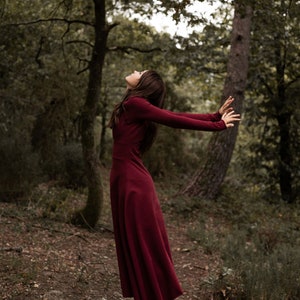 May include: A woman in a long, burgundy dress stands in a forest with her arms outstretched and head tilted back. She is wearing black and white sneakers with a red heart design.