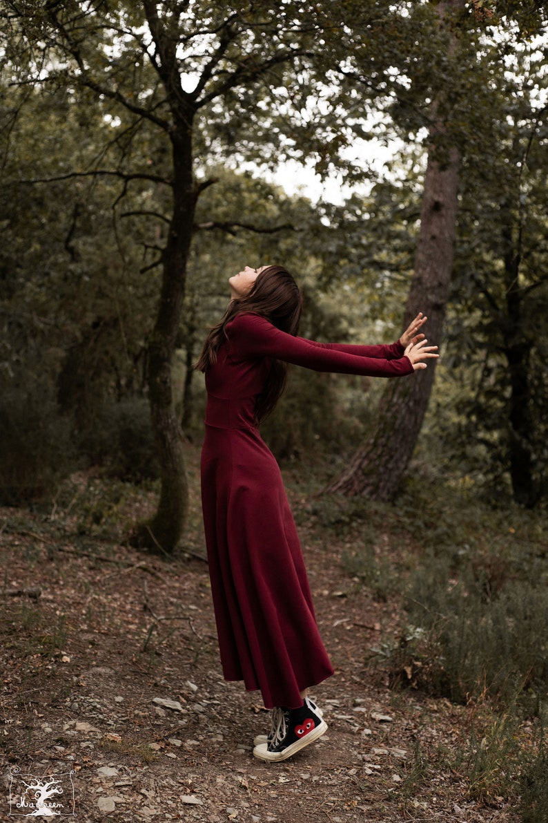 May include: A woman in a long, burgundy dress stands in a forest with her arms outstretched and head tilted back. She is wearing black and white sneakers with a red heart design.