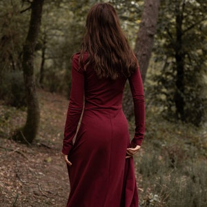 May include: A woman in a long, burgundy maxi dress walks through a forest. The dress has long sleeves and a fitted bodice. The woman's hair is long and brown.