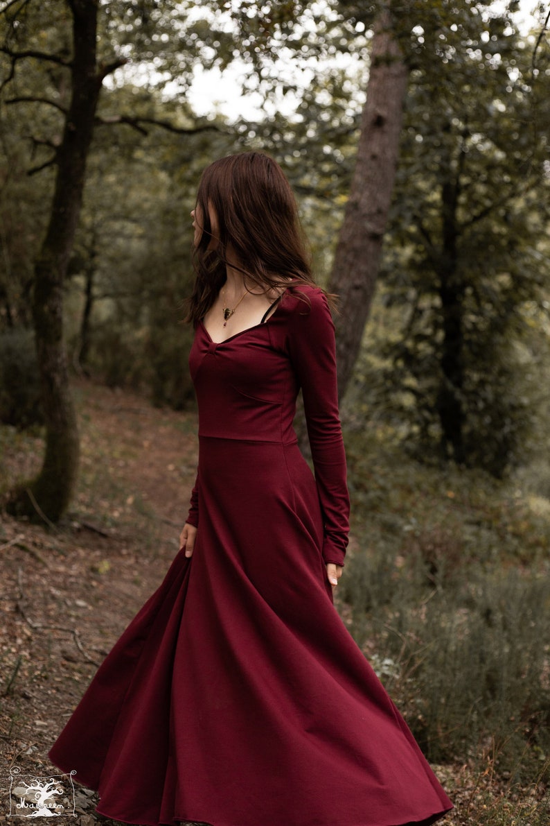 May include: A woman wearing a long-sleeved, burgundy maxi dress with a sweetheart neckline. She is standing in a forest setting with trees and foliage in the background.