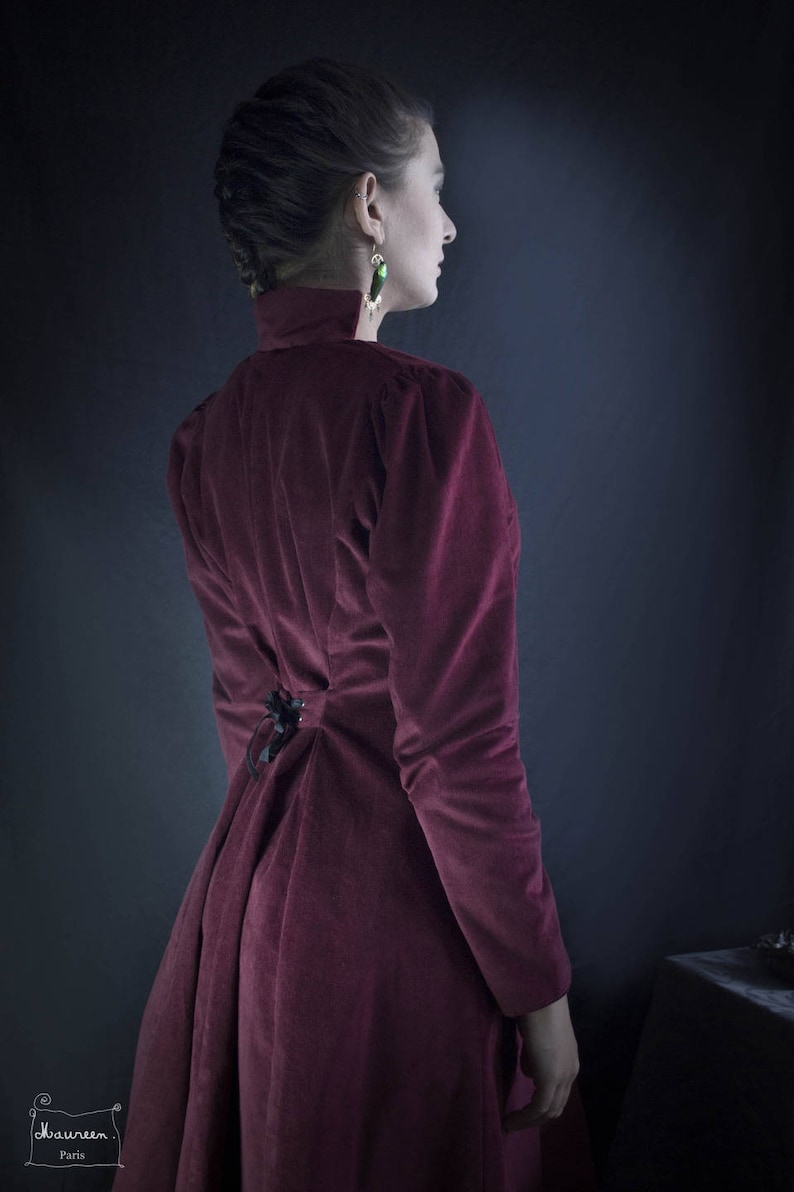 May include: A woman wearing a long, burgundy velvet dress with a lace-up back. The dress has a high neckline and long sleeves. She is wearing green earrings.