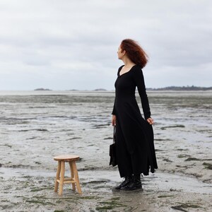 Pu&ograve; includere: Una donna con un vestito nero si trova su una spiaggia con uno sgabello di legno di fronte a lei. Sta guardando l'acqua.