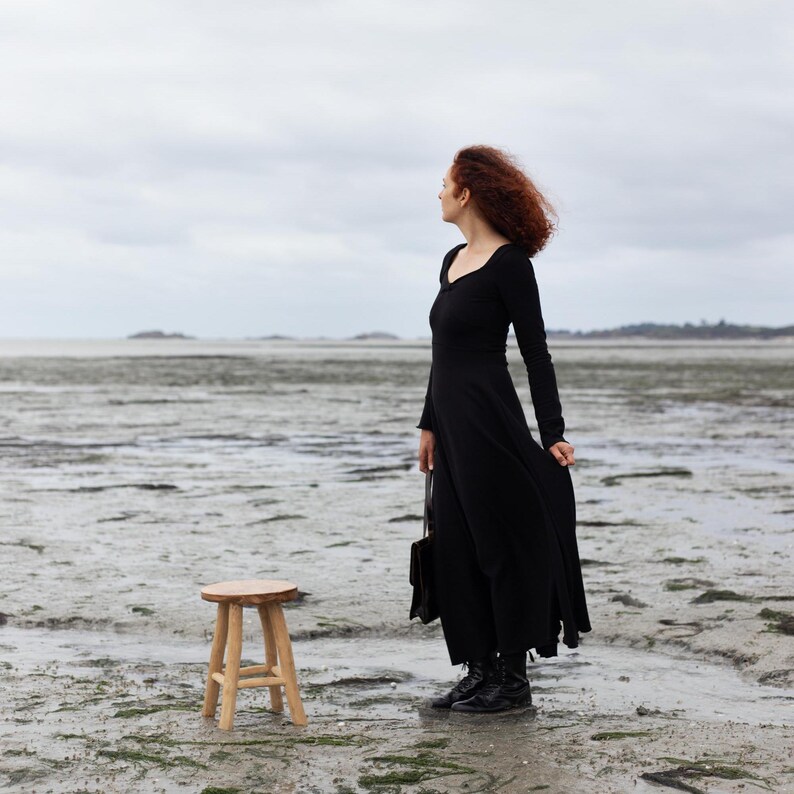 Pu&ograve; includere: Una donna con un vestito nero si trova su una spiaggia con uno sgabello di legno di fronte a lei. Sta guardando l'acqua.