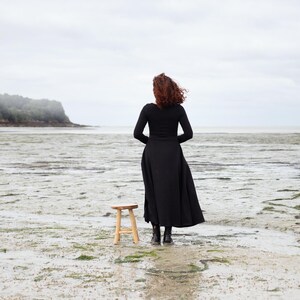 Pu&ograve; includere: Una donna con un vestito nero si trova su una spiaggia, di spalle alla telecamera, guardando l'oceano. Uno sgabello di legno si trova di fronte a lei.
