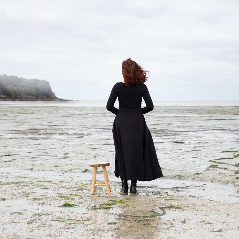 Pu&ograve; includere: Una donna con un vestito nero si trova su una spiaggia, di spalle alla telecamera, guardando l'oceano. Uno sgabello di legno si trova di fronte a lei.