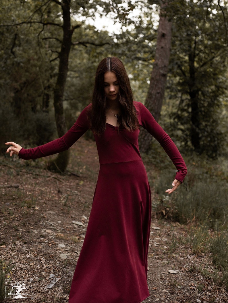 May include: A woman in a long, burgundy dress stands in a forest. The dress has long sleeves and a V-neckline. She is looking down at the ground.
