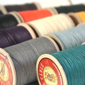 May include: Close-up of spools of colourful waxed cotton thread. The spools are arranged in a row, with the labels facing the viewer. The labels feature the text "Cable" and "50 grs".