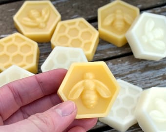 May include: A collection of hexagonal beeswax soap bars with different designs, including honeycombs and bees. The bars are in various shades of yellow and white.
