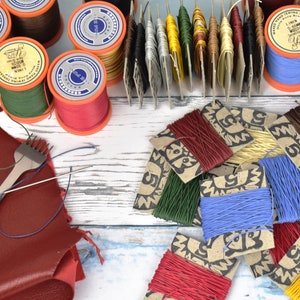 Peut inclure: Assortiment de bobines de fil de lin ciré de différentes couleurs, dont le rouge, le vert, le bleu et le brun. Les bobines sont disposées sur une surface en bois blanc avec un morceau de cuir brun et un poinçon à cuir. Le fil est emballé dans de petites boîtes en carton avec un motif imprimé.