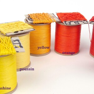 May include: A collection of colorful thread spools in shades of yellow, orange, and red. The spools are labeled with color names like "pistachio," "yellow," and "orange." The thread is wound on cardboard spools, ideal for crafts and sewing projects.