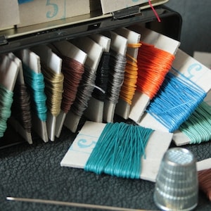 May include: A vintage sewing kit with spools of colourful thread in shades of blue, orange, brown, and green. The thread is wound on small cardboard bobbins. A silver thimble and a needle are also visible, suggesting sewing and crafts.