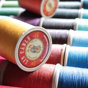 May include: A close-up of a collection of colourful spools of thread. The spools are arranged in a row, with the thread colours ranging from green to blue to red. The spools are labelled with the brand name "A.I.T. CABLE" and the text "50 GR. Fils TOULEMONDE AU CHINOIS".
