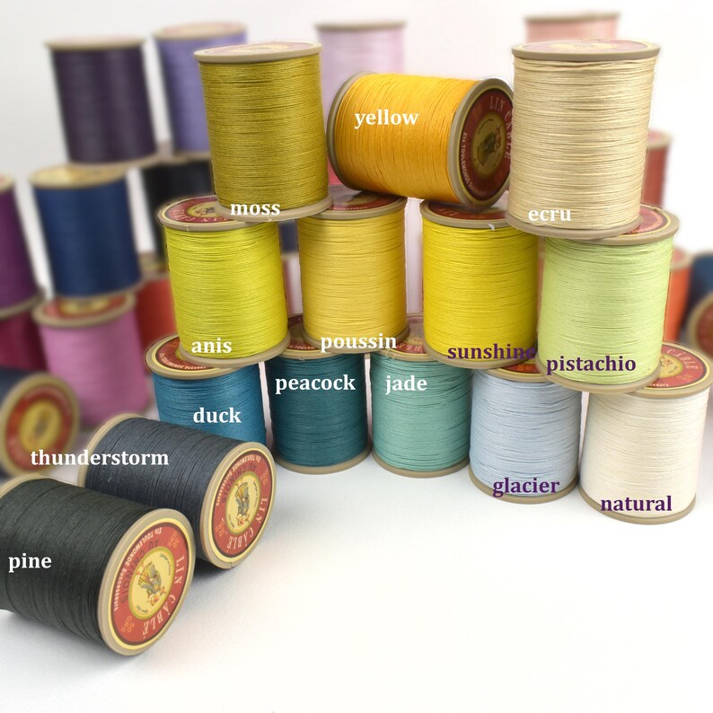 May include: A collection of colorful thread spools in various shades, including yellow, green, blue, and gray. Each spool is labeled with its color name, such as "moss," "yellow," and "ecru." The spools are arranged in a visually appealing stack, showcasing the range of colors.