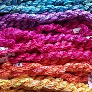 May include: A rainbow assortment of embroidery floss spools, with colors ranging from teal to yellow. The spools are labeled with the brand name "StrandSilk" and the size "12S".