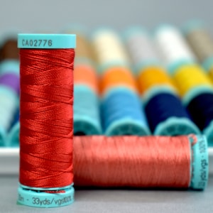 May include: A close-up of a red spool of thread with a turquoise label that reads "CA02776" and "33 yds/vgst00". The spool is sitting on a table with a row of other spools of thread in the background.