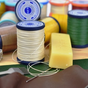 May include: Close-up of leatherworking tools and supplies, including spools of waxed thread in various colours, a bar of leather wax, a leather awl, and a piece of brown leather.