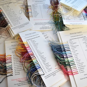 May include: A close-up of three colour charts for embroidery floss. The charts are labelled with the names of the colours and their corresponding numbers. The floss is arranged in rows by colour, with the ends of the floss hanging down.