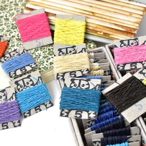May include: A selection of colourful thread spools in shades of pink, yellow, orange, blue, and green. The spools are on small cardboard cards with a panda design and the number 5. Some are in a divided box, others are scattered around.