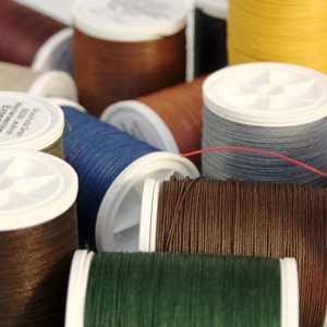 May include: Assortment of spools of waxed thread in various colours, including brown, red, green, blue, and beige. The spools are labelled with the brand name "Saphir" and the text "Epoxi-Waxed Thread".