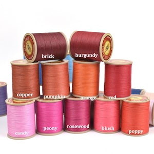 May include: A collection of colorful spools of thread, each labeled with a different color name. Colors include brick, burgundy, copper, pumpkin, orange, red, candy, peony, rosewood, blush, and poppy. The spools are arranged in a visually appealing display.
