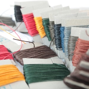 May include: A selection of colourful waxed thread spools in shades like red, yellow, green, and blue. The spools are arranged on cardboard holders, with some threads unwound. The image displays a variety of colours and textures, suitable for craft projects and sewing.