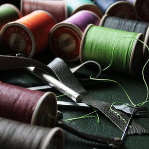 May include: A collection of colorful thread spools in shades of green, orange, purple, and burgundy. Leather strips and a stitching tool are also visible, suggesting a craft or DIY project. The spools are arranged on a dark surface.