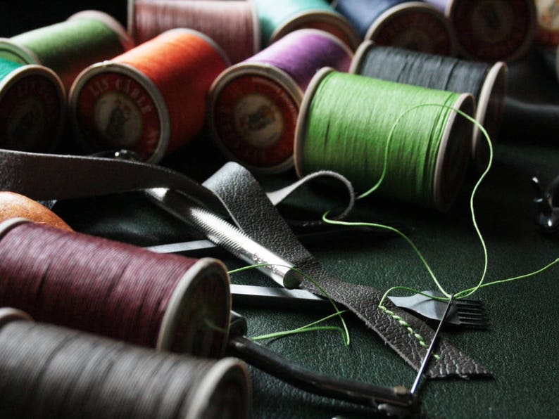 May include: A collection of colorful thread spools in shades of green, orange, purple, and burgundy. Leather strips and a stitching tool are also visible, suggesting a craft or DIY project. The spools are arranged on a dark surface.