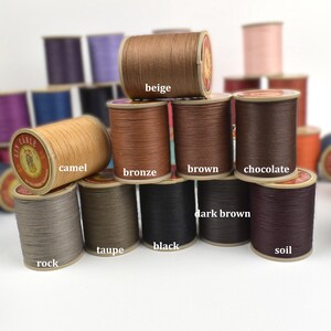 May include: A collection of spools of thread in various colors, including beige, camel, bronze, brown, chocolate, dark brown, black, and soil. The spools are labeled with their respective color names.