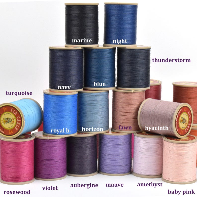 May include: A collection of spools of thread in various colors, including marine, night, navy, blue, turquoise, royal blue, horizon, fawn, hyacinth, rosewood, violet, aubergine, mauve, amethyst, and baby pink. Each spool is labeled with its color name.