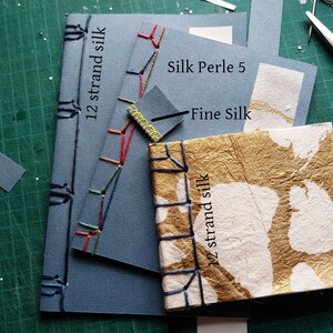 May include: Three different types of thread used for bookbinding. The first is a blue bookbound with 12 strand silk thread. The second is a blue bookbound with Silk Perle 5 thread. The third is a white and gold bookbound with 12 strand silk thread. The text "Fine Silk" is written on the image.