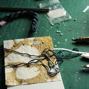 May include: A close-up of a handmade paper card with gold leaf accents. A needle and thread are used to stitch the card together. The card is on a green surface.