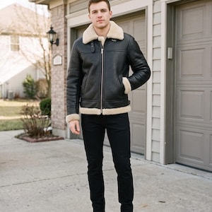 May include: A black leather jacket with a cream-colored shearling collar, cuffs, and hem, featuring a front zipper closure, side pockets, and adjustable side buckles, paired with black trousers and black leather boots.