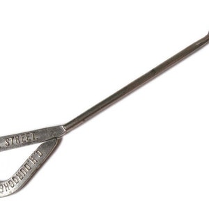 May include: A vintage metal button hook featuring a long, slender handle and a curved hook at the end. The teardrop-shaped handle is embossed with the text BANK STREET and C.H. DUROCHER & CO. The tool has a weathered, metallic grey finish.